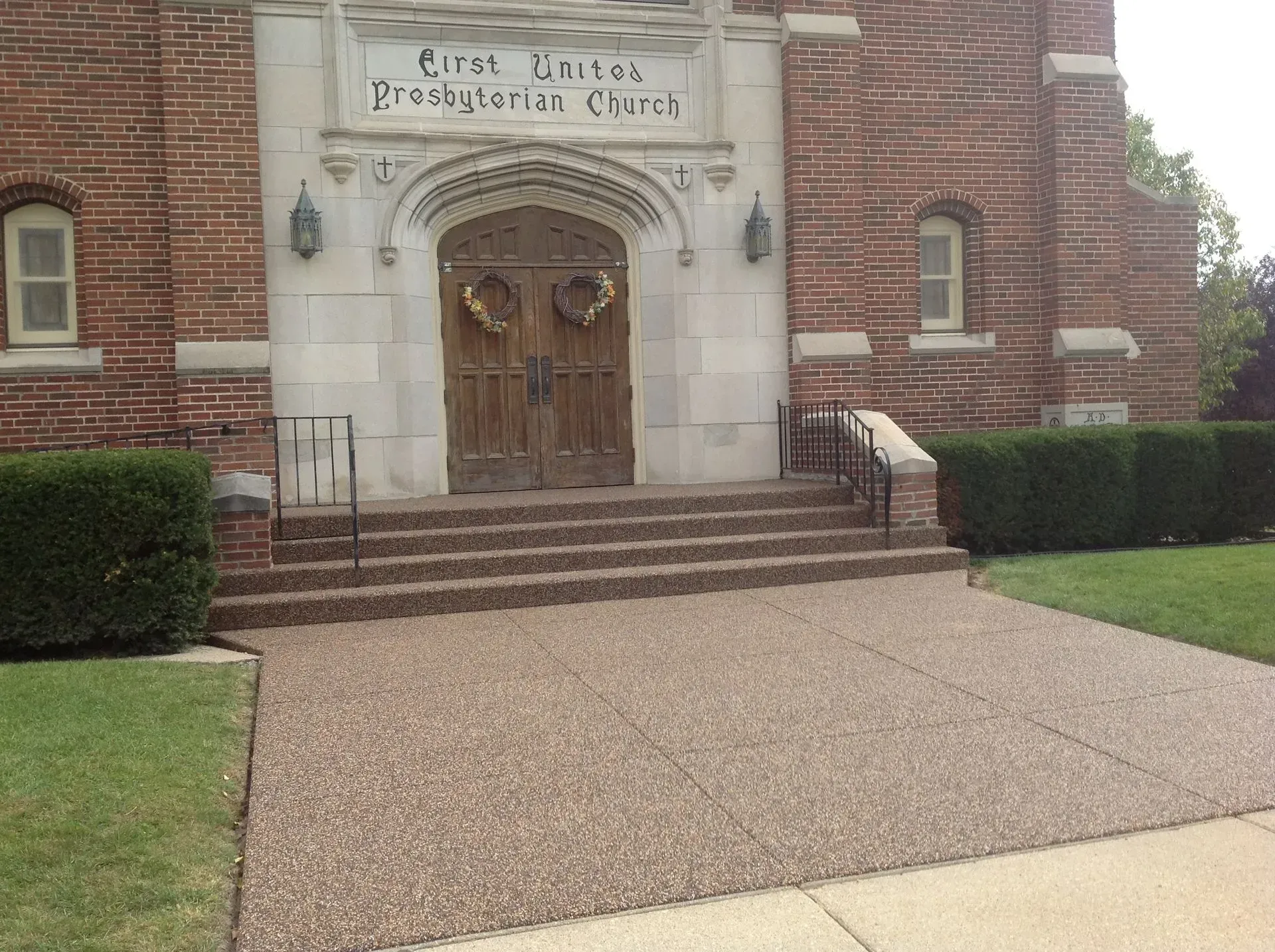 Brick front entrance of First United Presbyterian Church with wooden double doors, steps, and a pebbled concrete walkway.