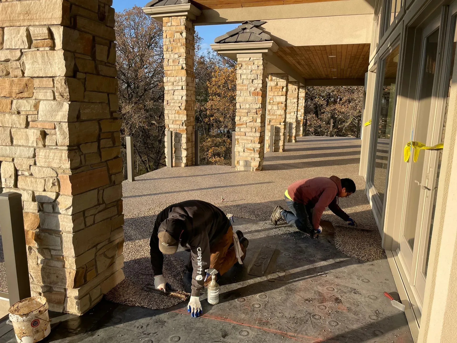 Two people kneeling on a patio under a stone-pillared walkway, working on the gravel surface near a building.
