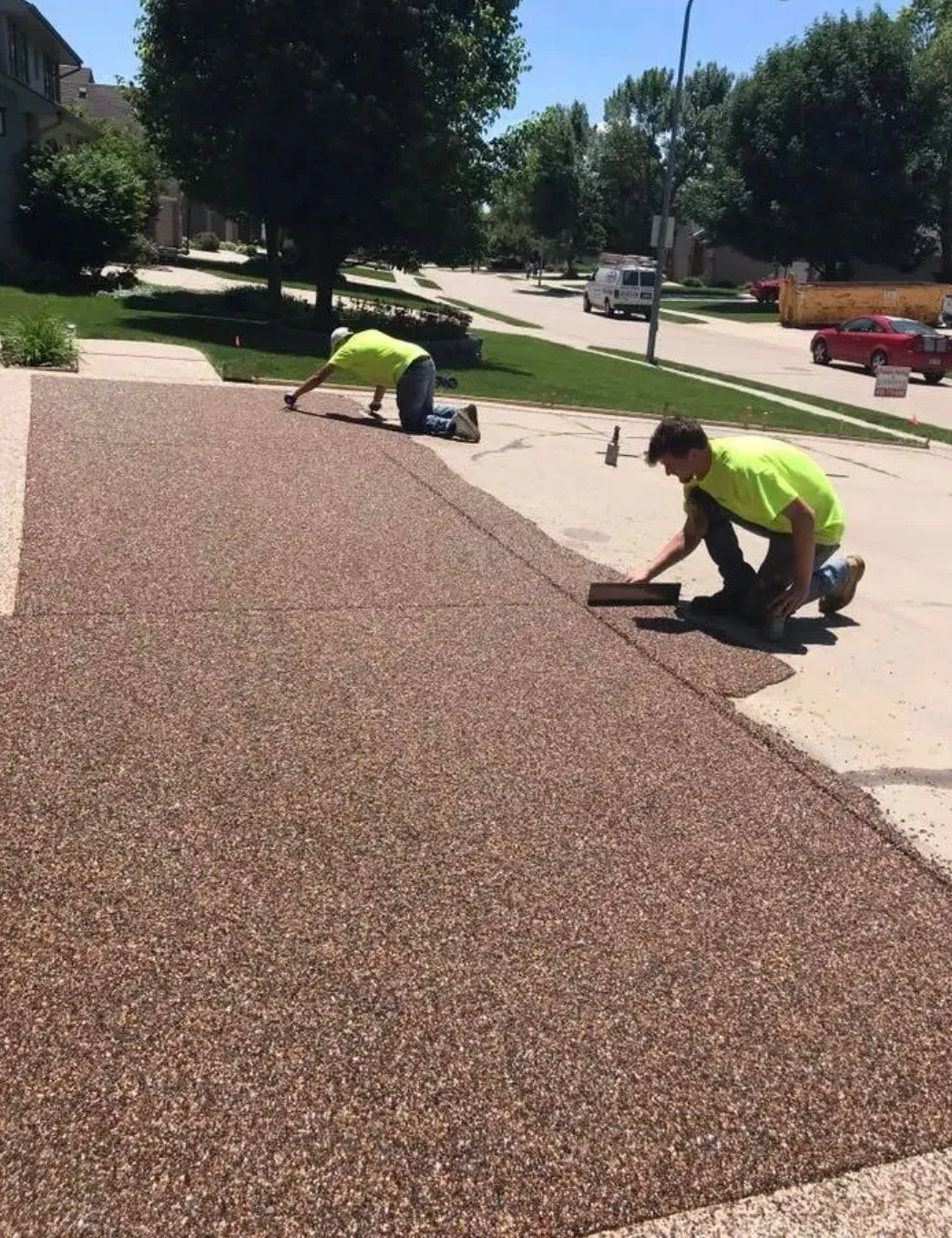 Two people in neon yellow shirts kneeling on a driveway, applying a textured, brown resin-based stone coating.
