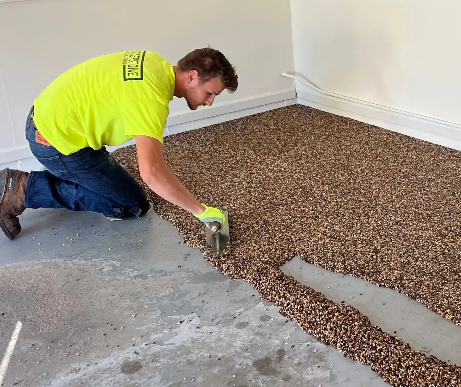 A person in a neon yellow shirt kneeling to apply a layer of small brown pebbles over a gray floor surface.