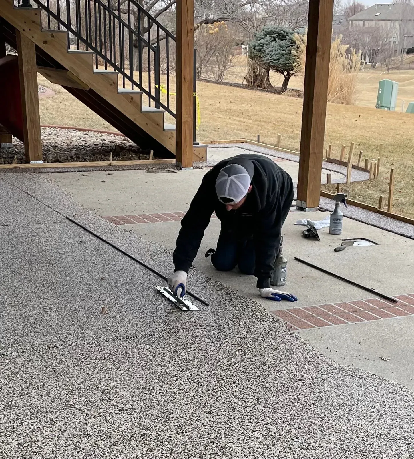 A worker kneeling on a concrete patio, using a trowel to install black divider strips into a new stone-texture overlay.