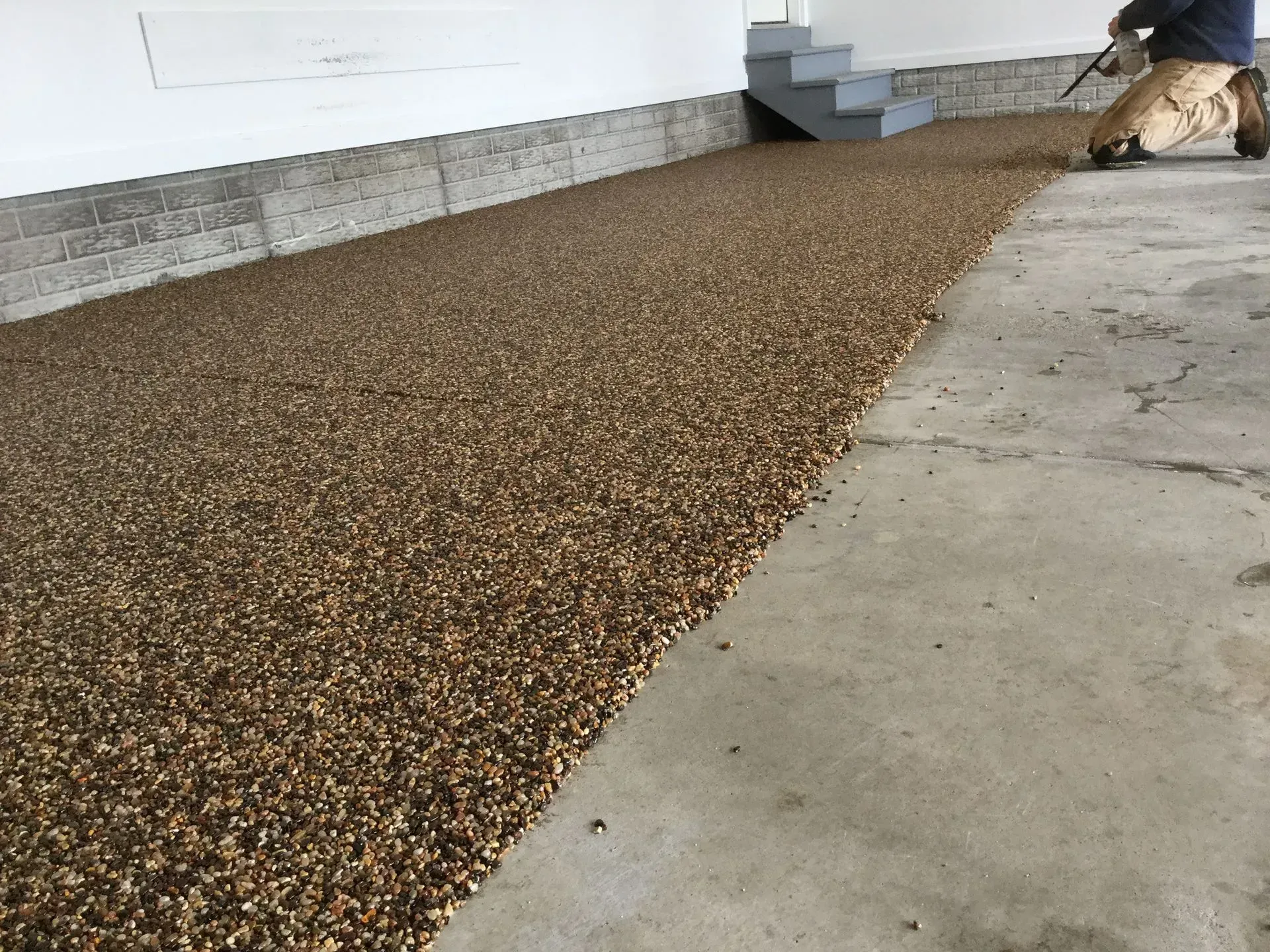 A person applies brown, textured epoxy aggregate coating to a concrete garage floor near a set of stairs.