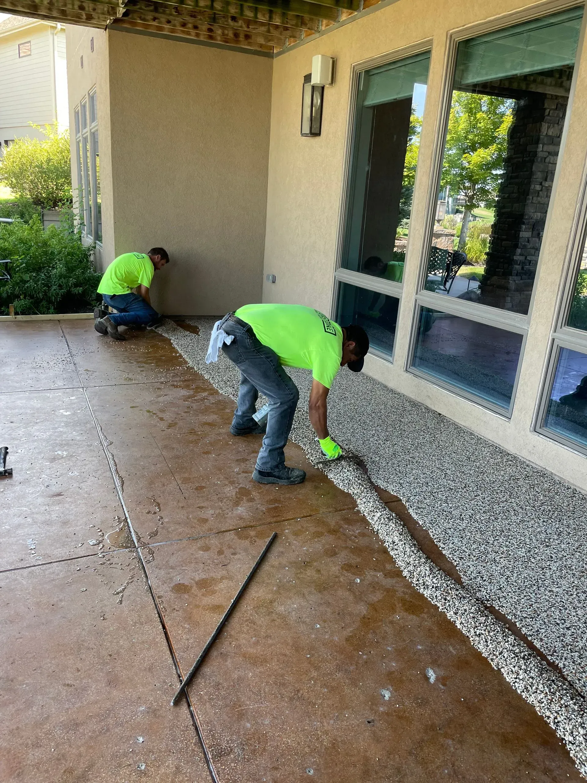 Two workers in neon yellow shirts install gravel edging along the foundation of a house patio.