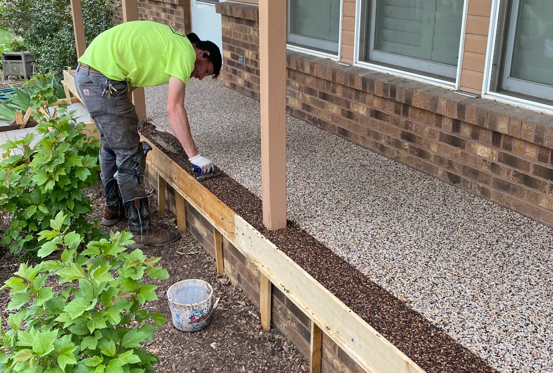 A person in a neon yellow shirt works on a wooden landscaping border in front of a brick house with gravel ground.