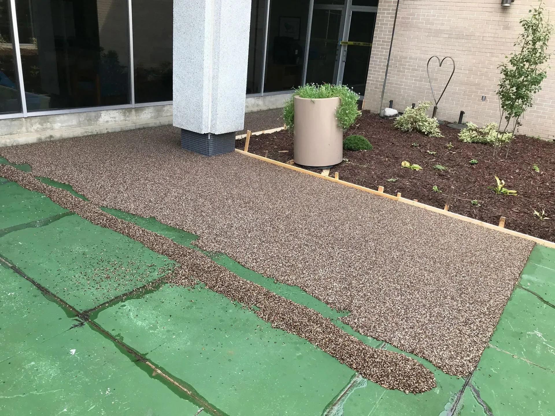 A section of a green, paved ground partially covered by loose brown gravel near a light-colored pillar and garden bed.