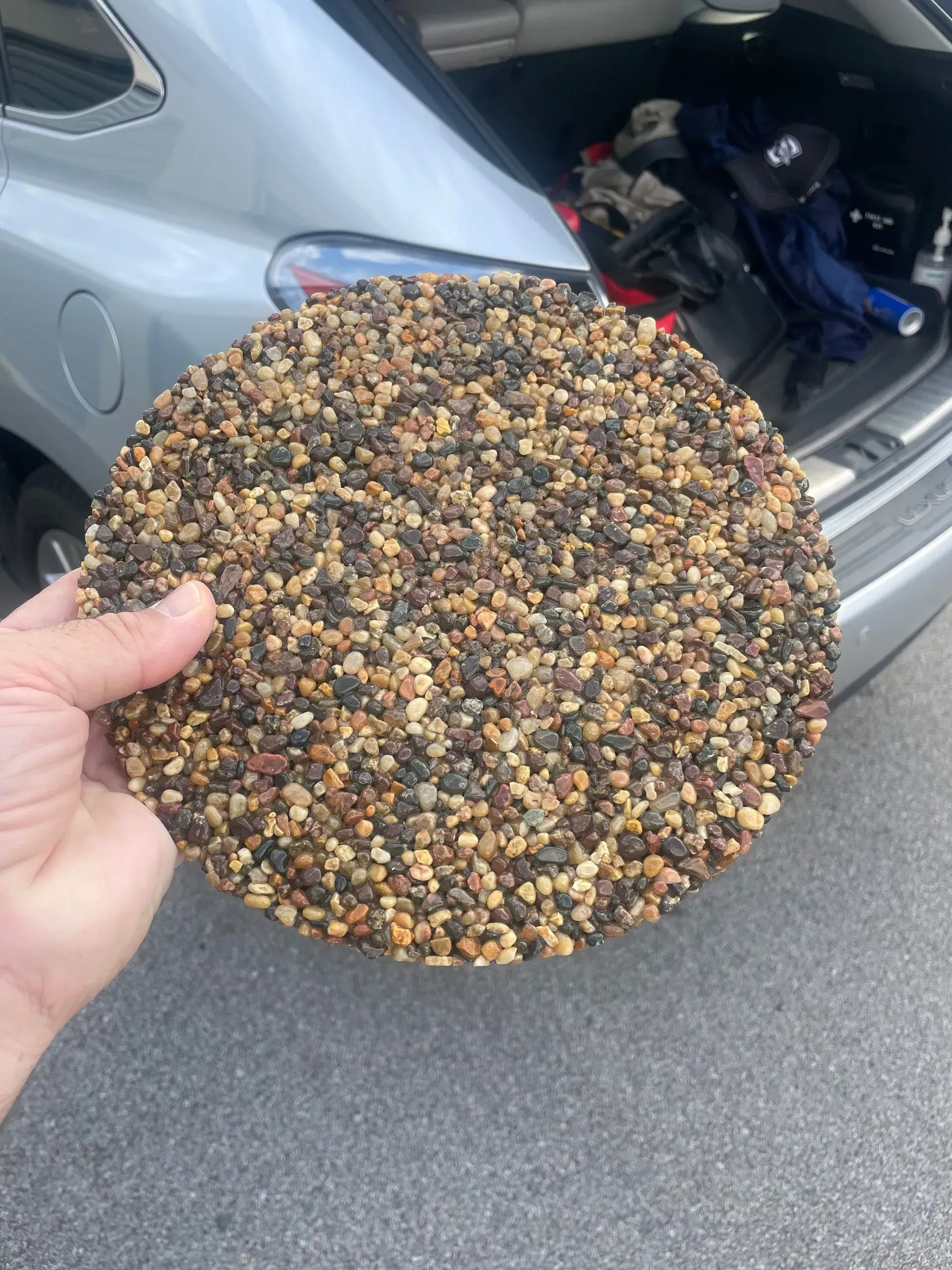 A hand holds a circular pad covered in a textured mosaic of small, multicolored tan and dark brown pebbles.