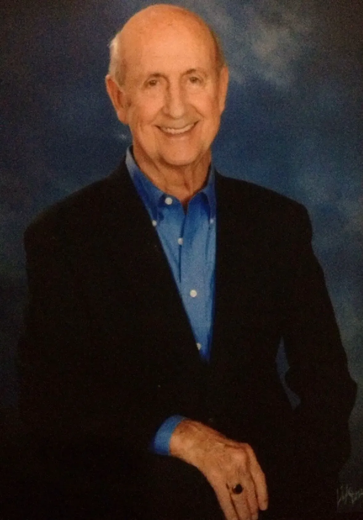 A smiling individual wearing a dark blazer over a blue button-down shirt, posed against a blue, mottled background.