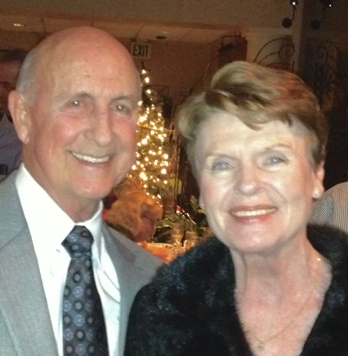 A smiling couple poses for a photo at an indoor event, with a lit Christmas tree in the background.