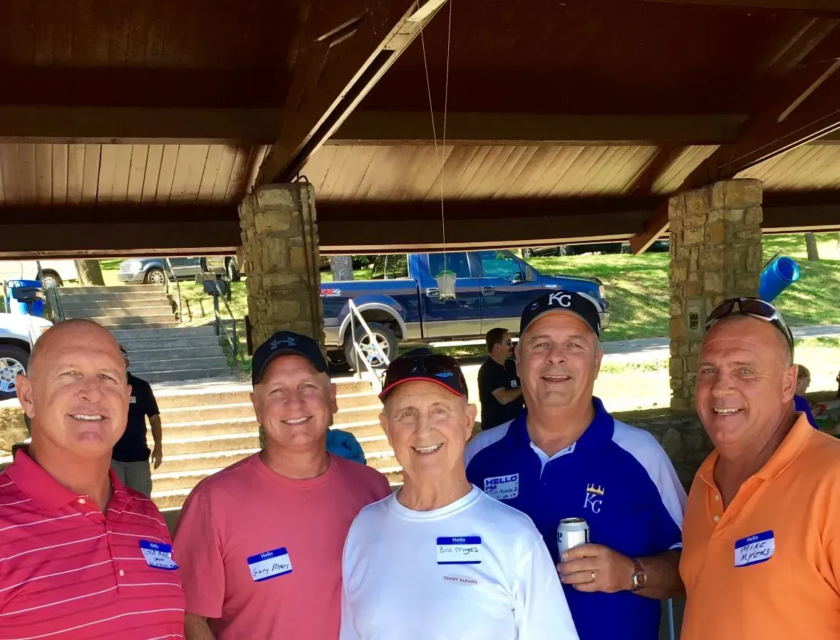 Five people posing for a photo under an open-air pavilion during a daytime gathering, wearing casual shirts and name tags.