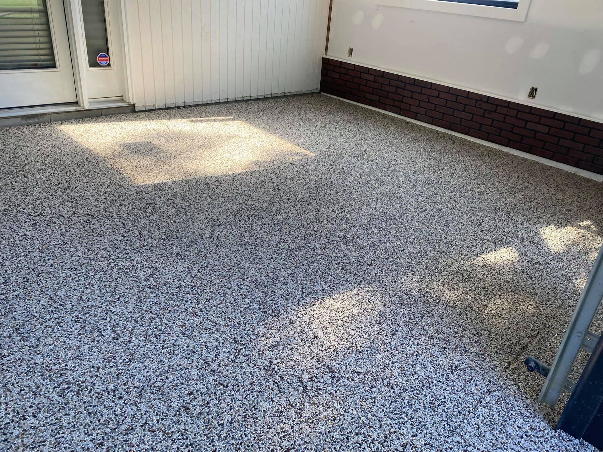 An outdoor patio with a speckled gray and white epoxy floor, light-colored vertical siding, and dark brick trim