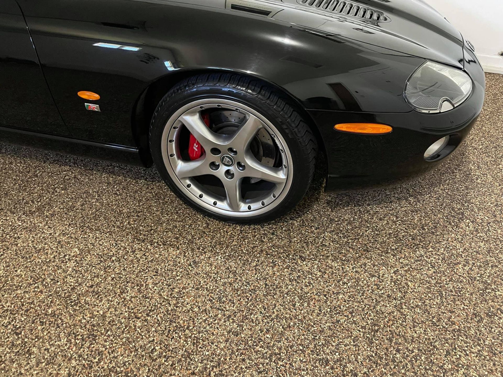 A black Jaguar sports car, showing the front passenger side wheel and fender, parked on a speckled garage floor