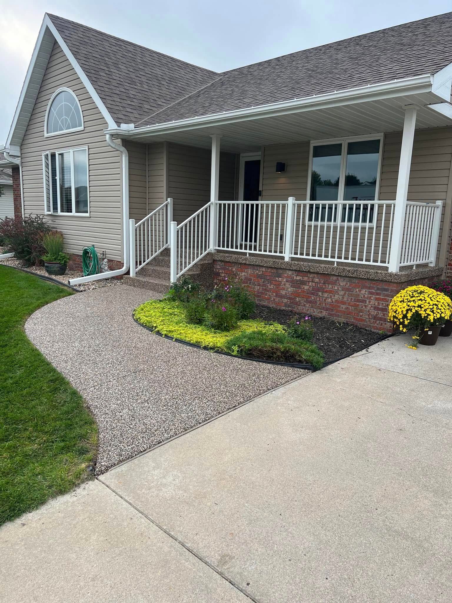 A taupe-sided house with a brick foundation, covered porch with white railing, and a gravel walkway leading to the entry