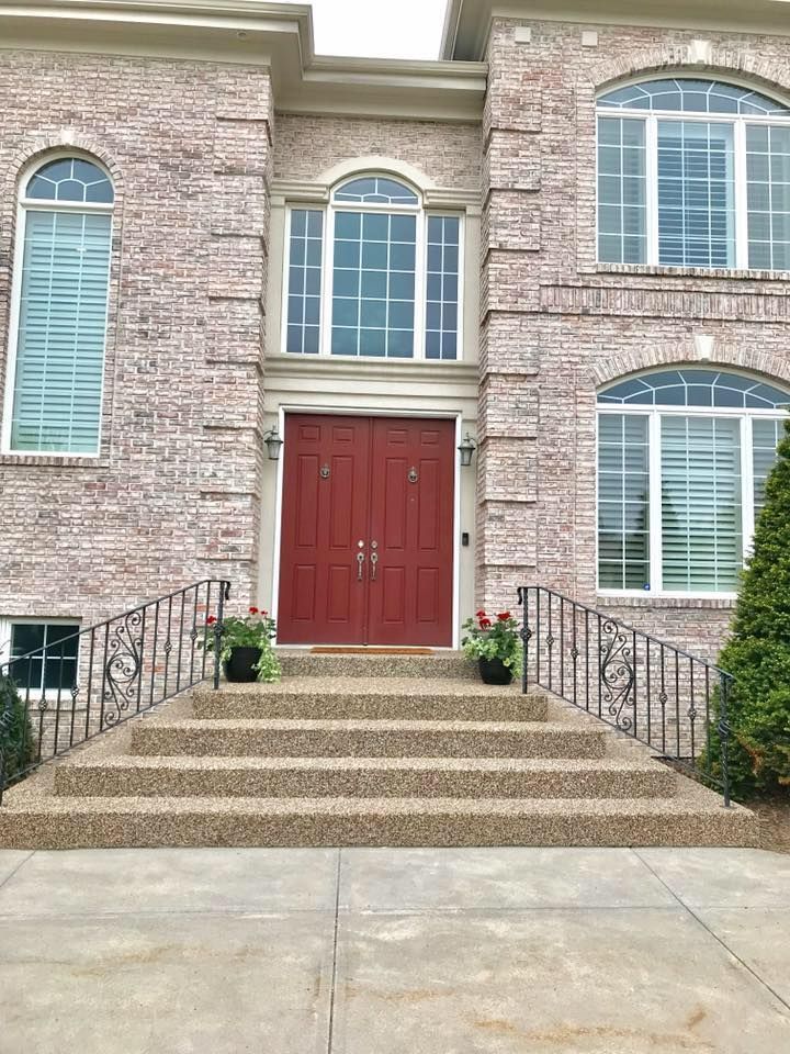 A two-story brick house with a red double front door, concrete steps, iron railings, and arched windows