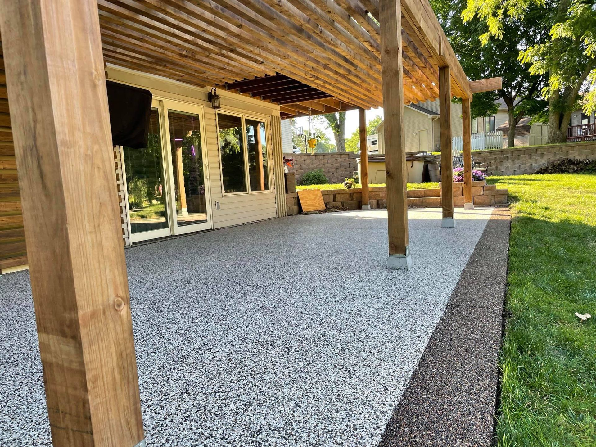 A patio with a speckled light gray surface and dark brown border, shaded by a wooden pergola attached to a house