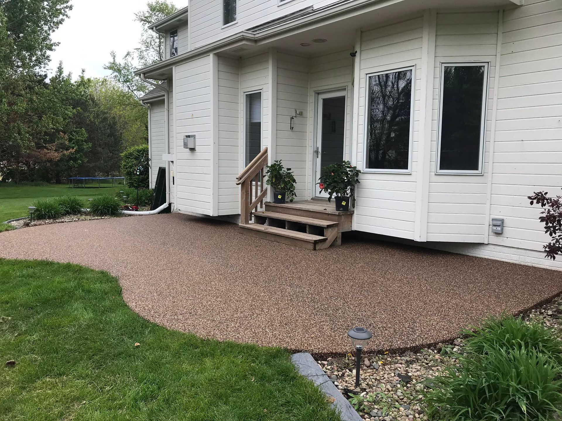 A house exterior features a textured pebble patio with small wooden stairs leading to a white door next to bay windows