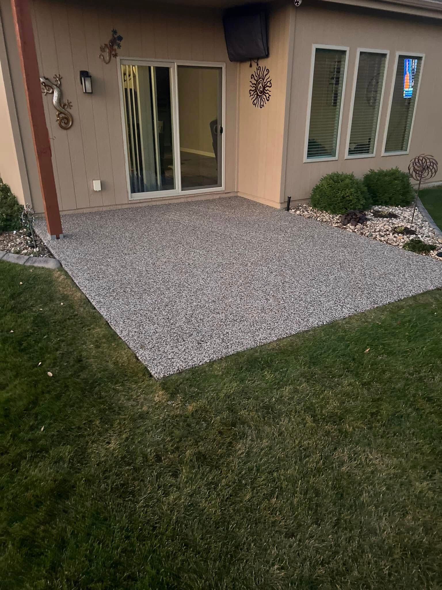A rectangular patio made of small gray decorative stones sits outside a house, next to a sliding glass door and lawn