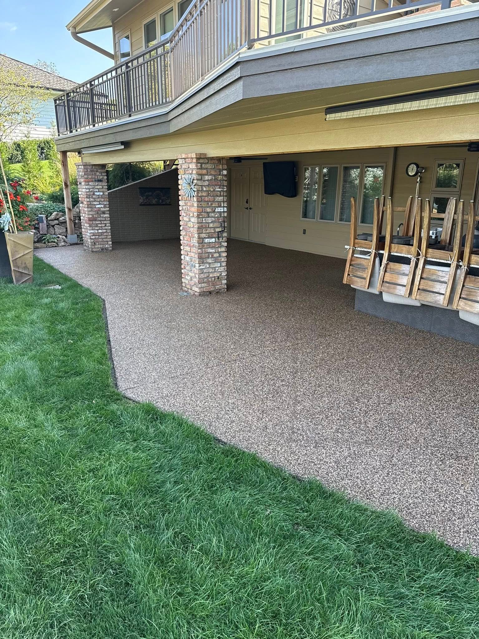 A two-story house with a beige exterior, a pebble-textured patio area, brick support pillars, and a green lawn