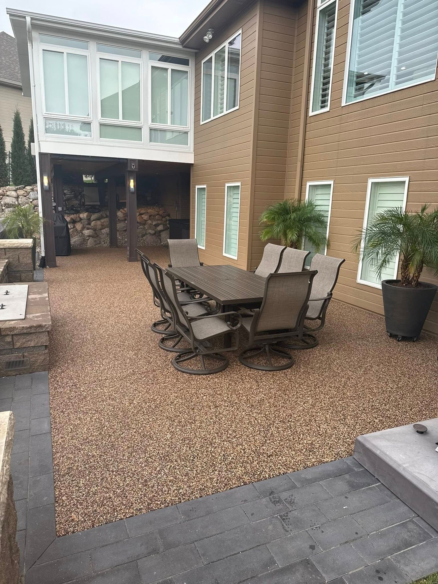 A tan house exterior with a gravel patio, outdoor dining set, and potted plants under a white upper deck