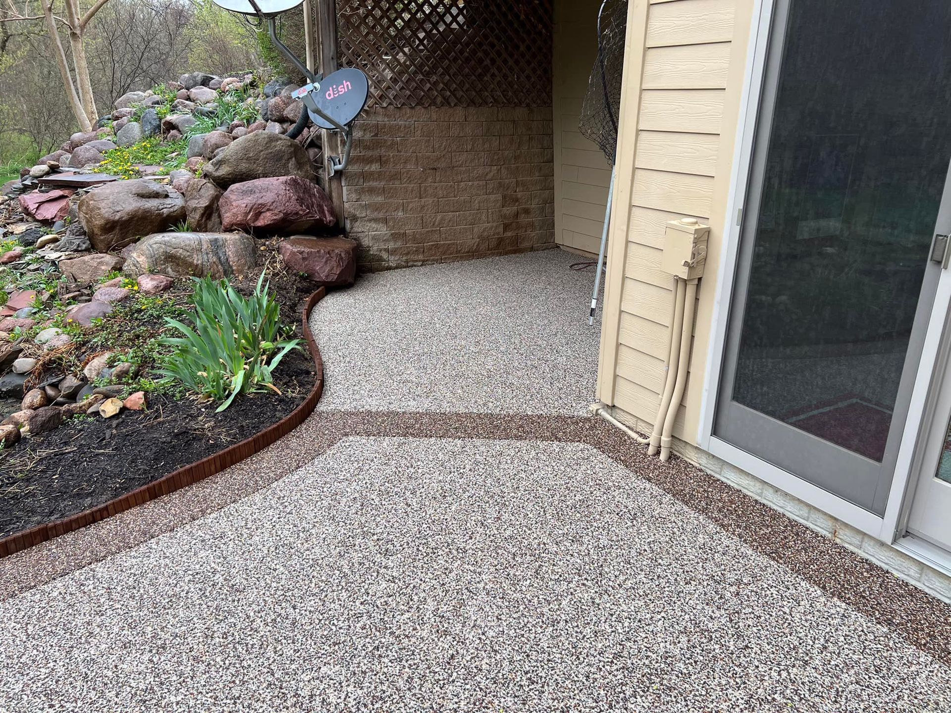 A ground-level patio made of light, multi-colored aggregate concrete with a dark border, next to a sliding glass door