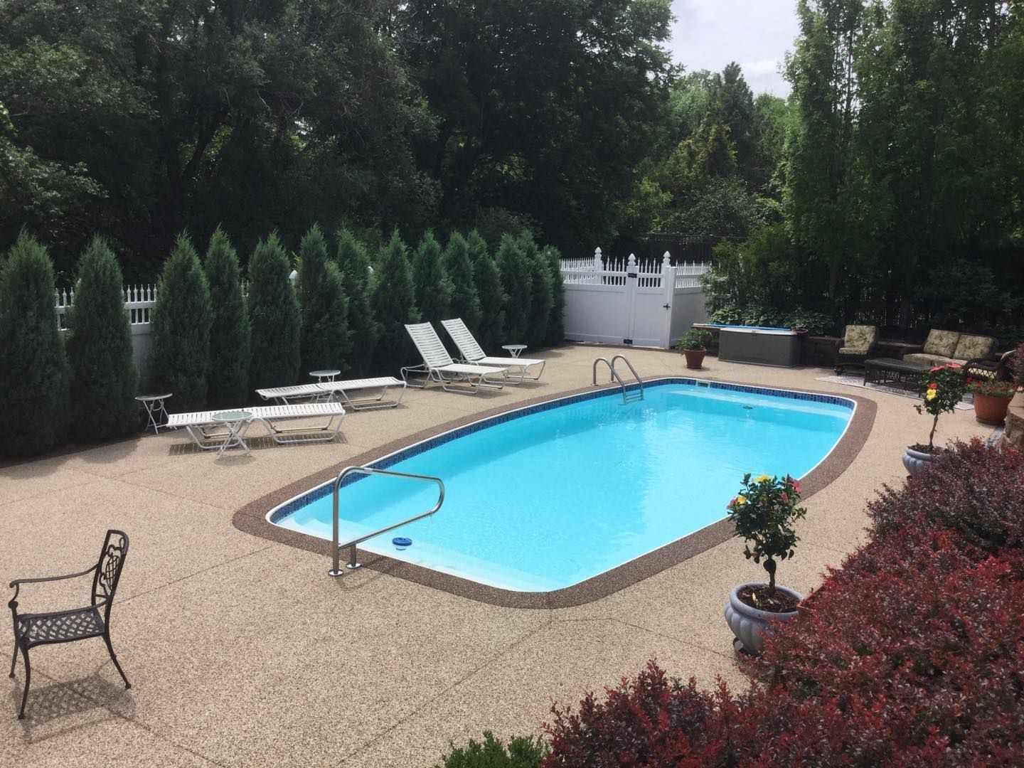 An outdoor rectangular swimming pool with blue water, surrounded by patio furniture, manicured hedges, and a white fence