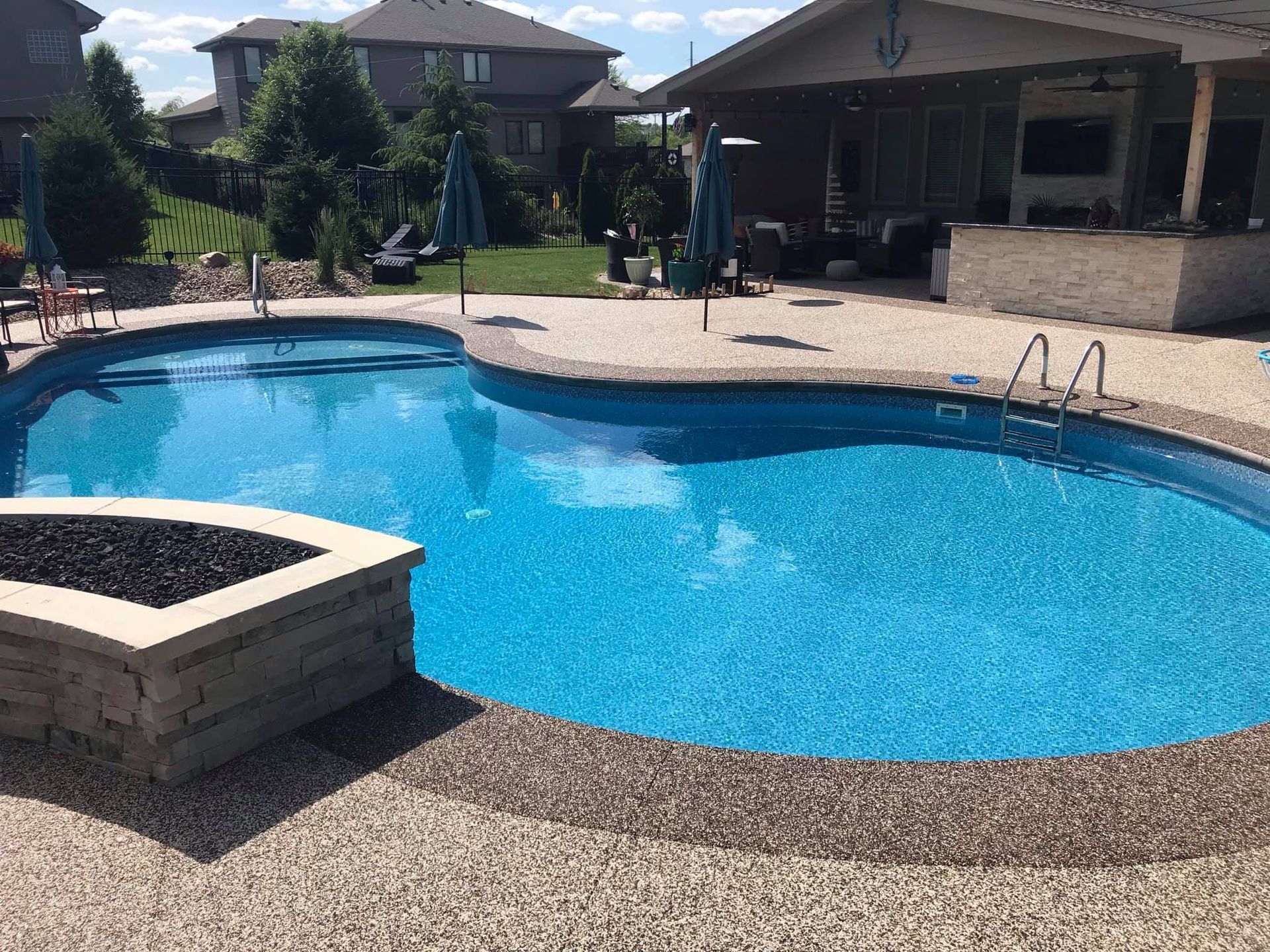 A kidney-shaped swimming pool with a stone fire pit in the foreground, set in a sunny backyard patio area
