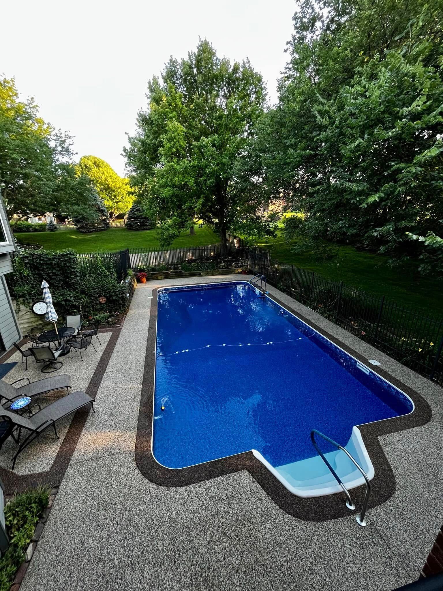 An elevated view of a bright blue backyard swimming pool surrounded by a light-colored gravel patio and lush green trees