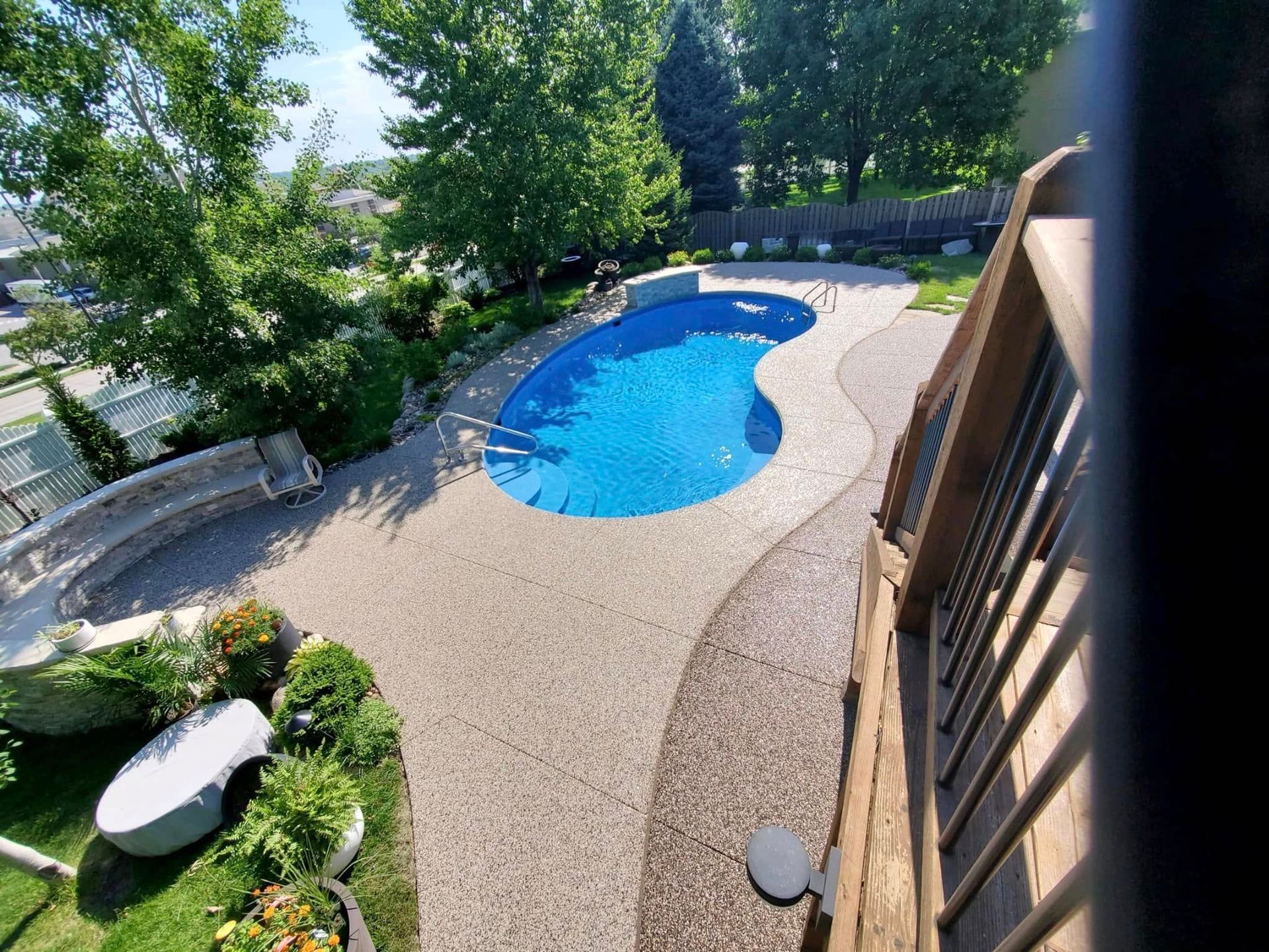 An aerial view of a bright blue kidney-shaped swimming pool surrounded by a light-colored pebbled patio and green trees