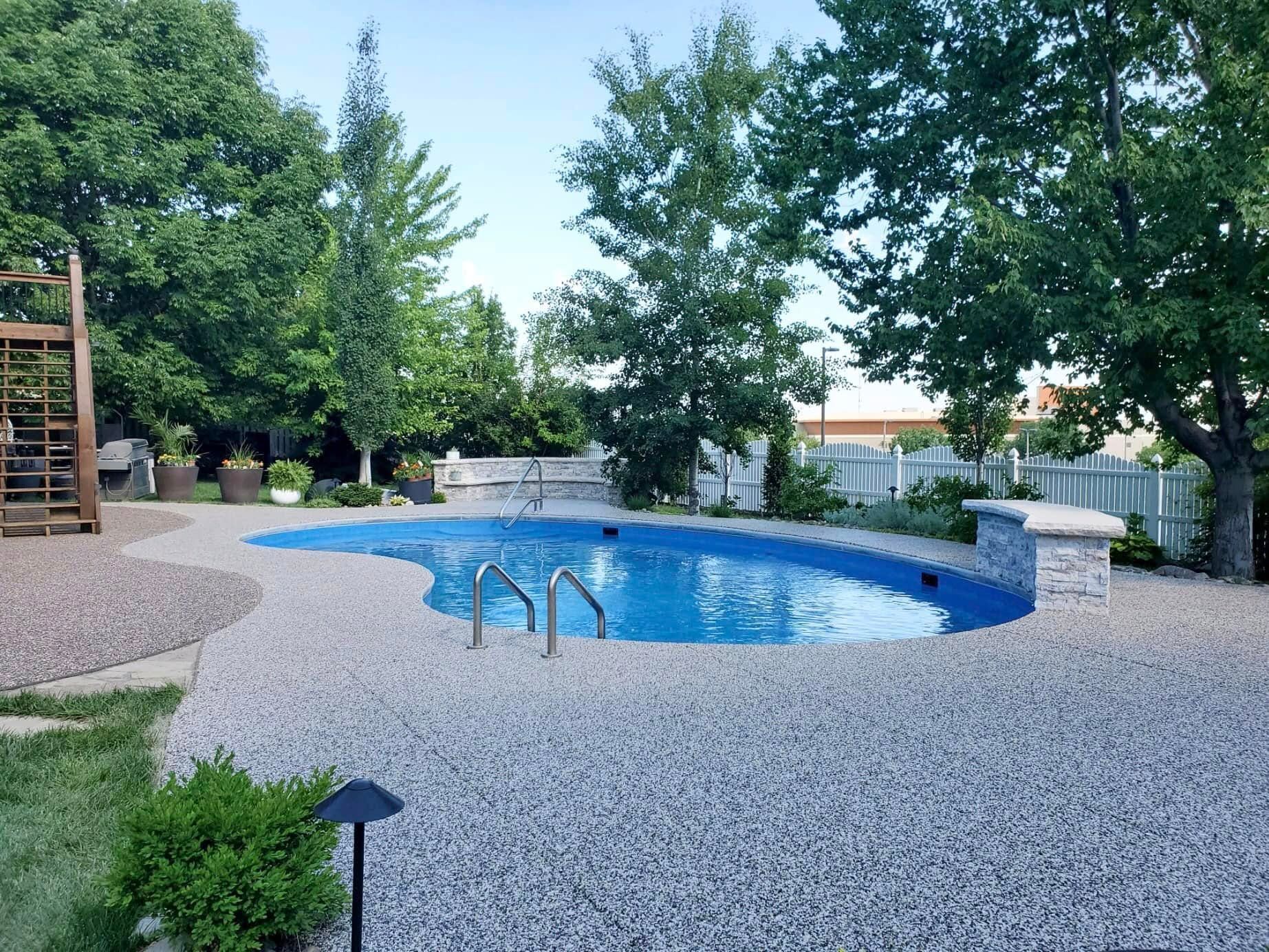 A kidney-shaped swimming pool in a backyard, surrounded by light-colored gravel, trees, and a wooden play structure