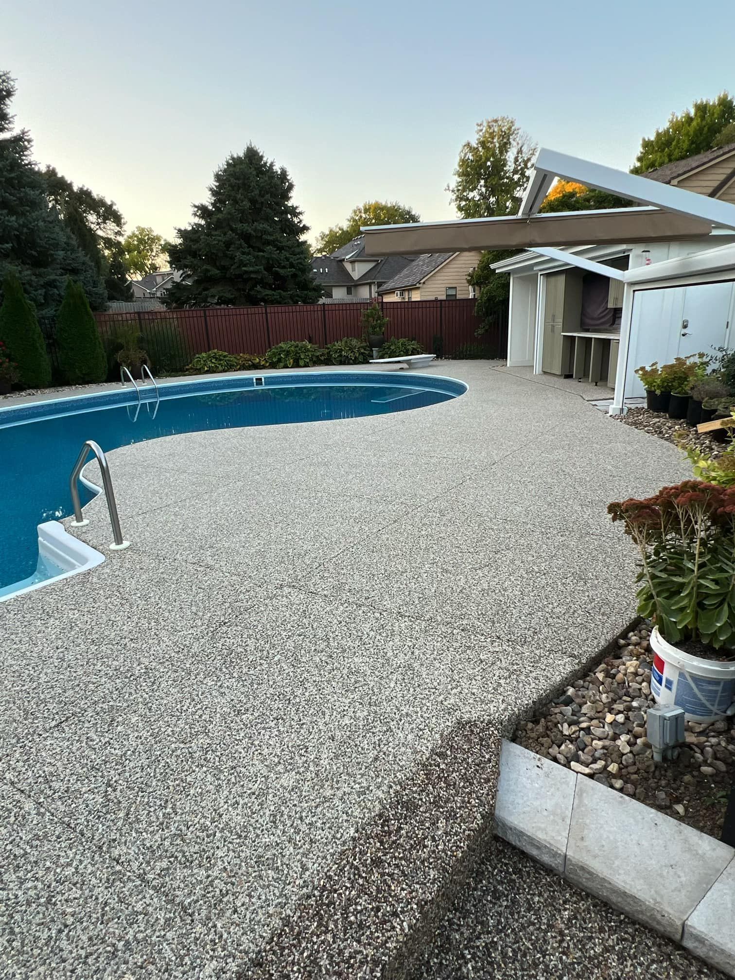 A backyard pool with a textured gravel deck, a white pergola structure, and surrounding trees at sunset