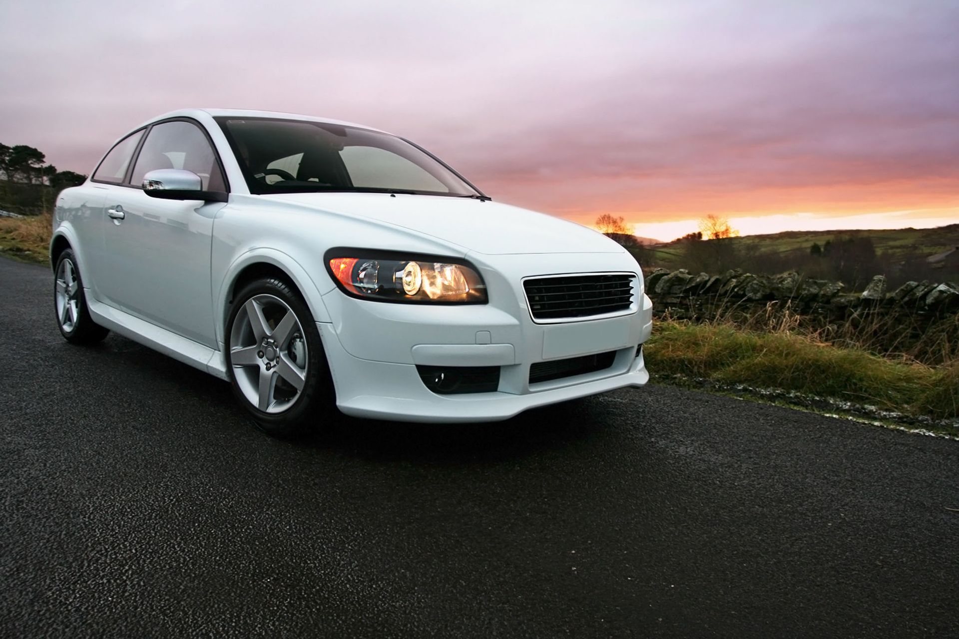 White Volvo C30 coupe parked on a wet road at sunset.