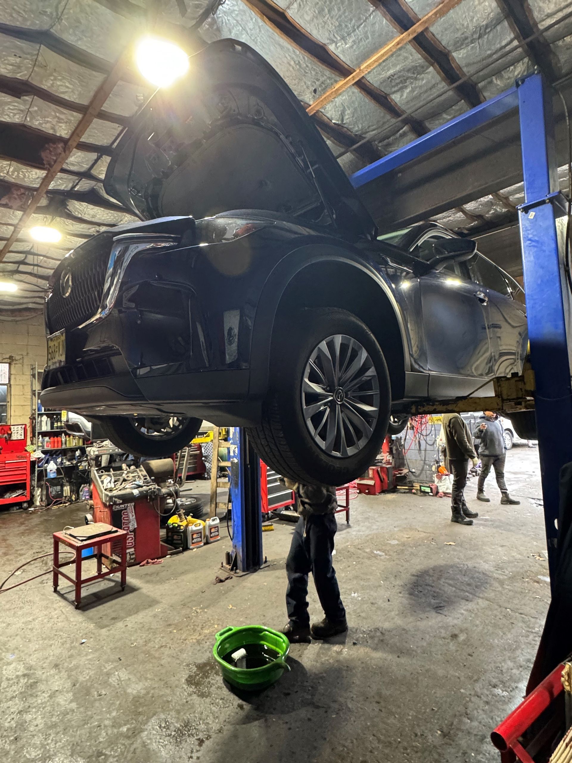 Car on a lift in a garage with hood open, mechanic draining fluid into a green basin.