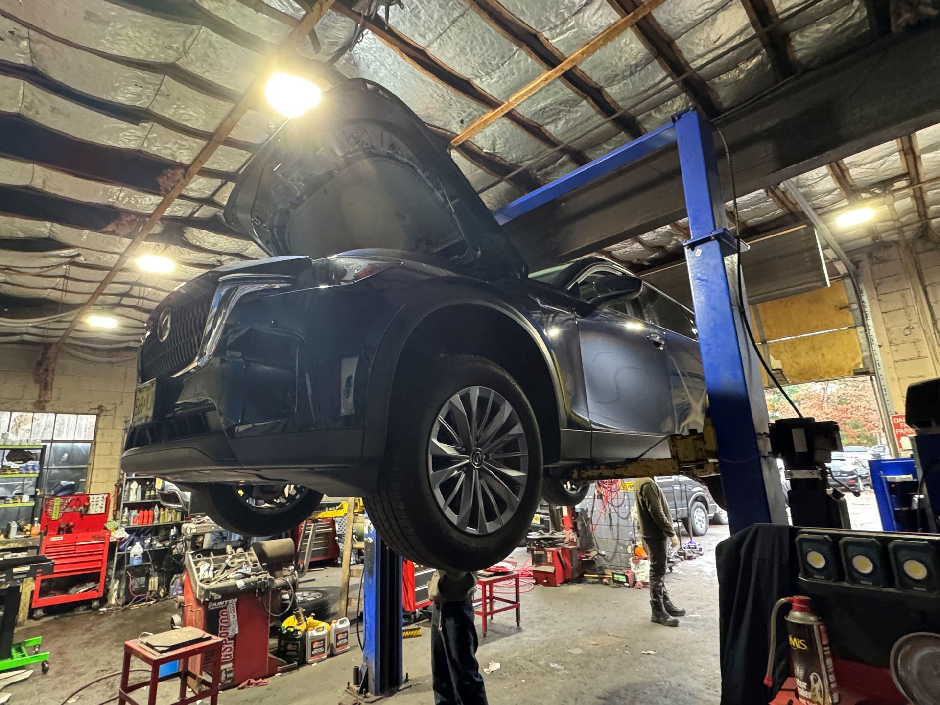 Car on a lift in a repair shop with hood open, under bright lights. A mechanic stands nearby.