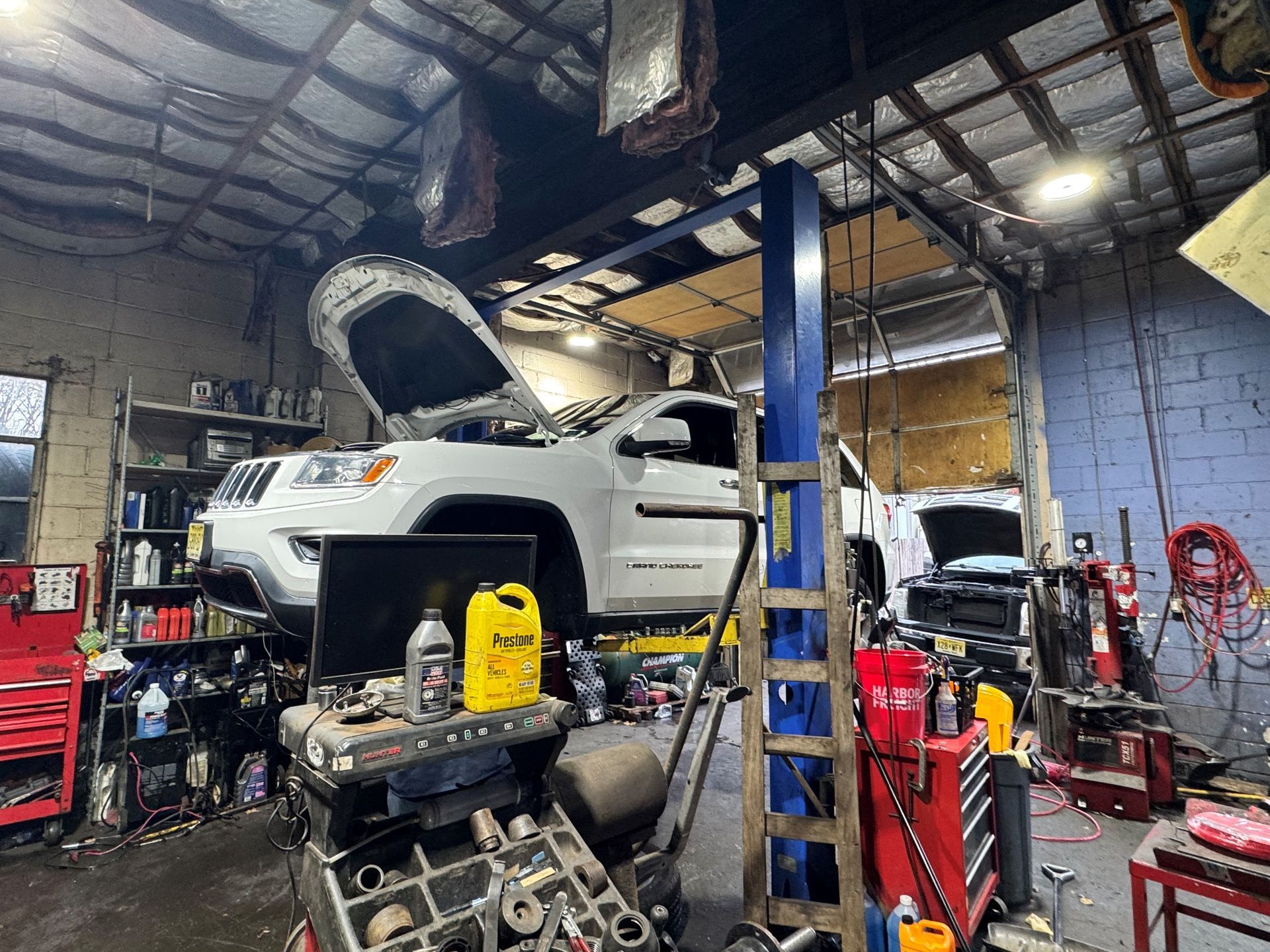 White Jeep on a lift in a cluttered auto repair shop, hood open. Tools and equipment surround it.