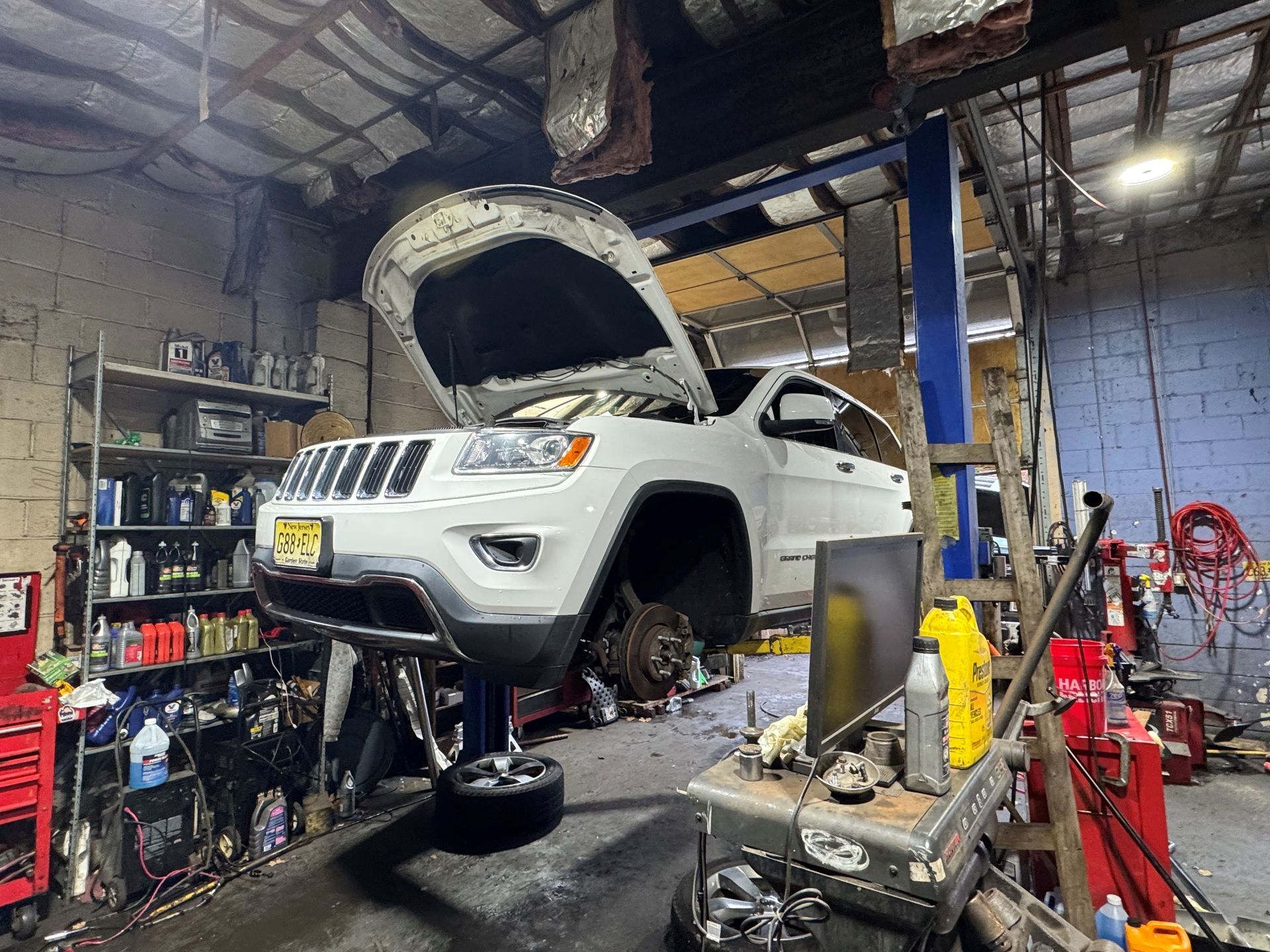 White Jeep Grand Cherokee raised in a repair shop with hood open, wheels removed.