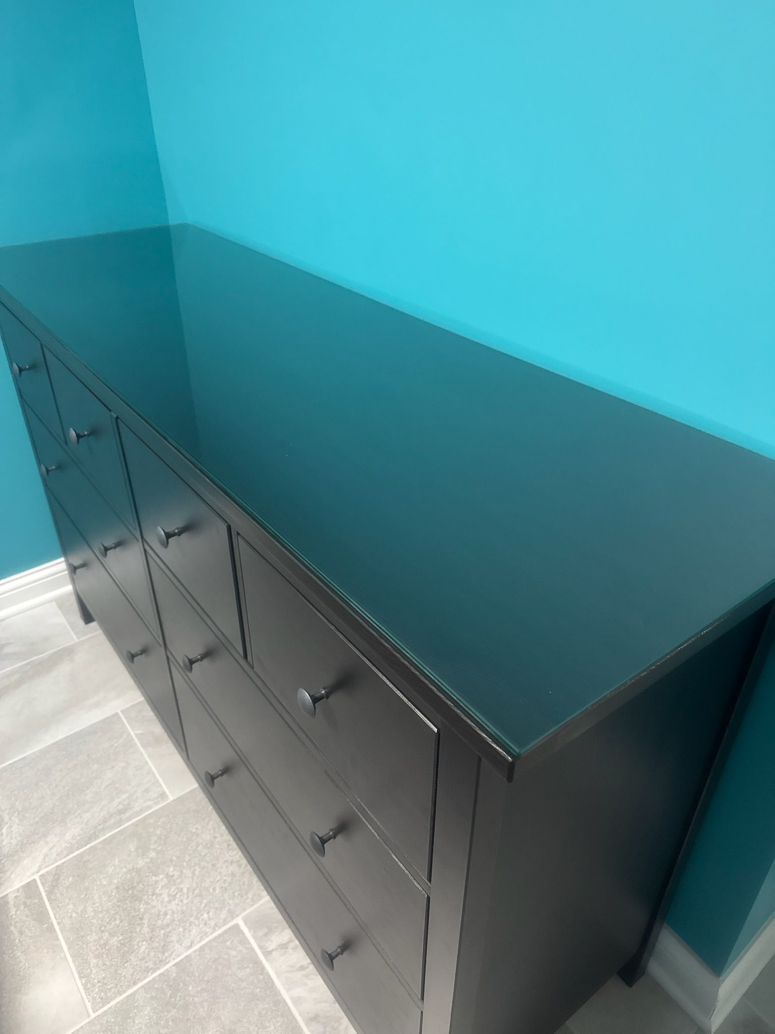 A high-angle view of a dark dresser with glass top, positioned against a vibrant turquoise wall and tile floor.