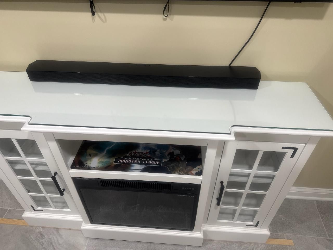 A white fireplace console with a glass top and a black soundbar, positioned against a beige wall.