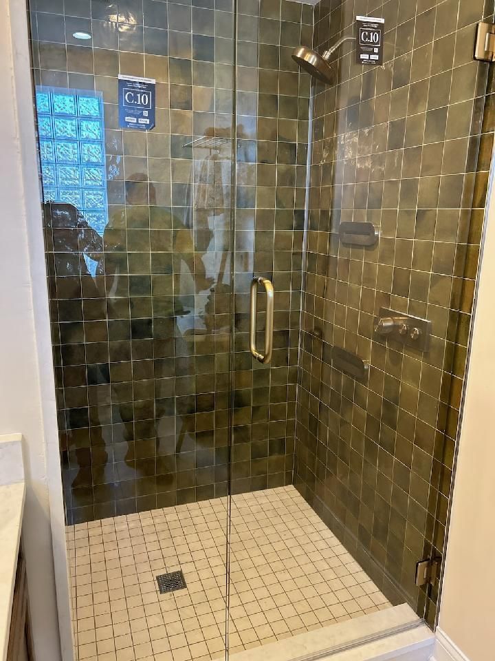 A modern shower with dark, square-tiled walls, a glass door with a gold handle, and a white, tiled shower floor.
