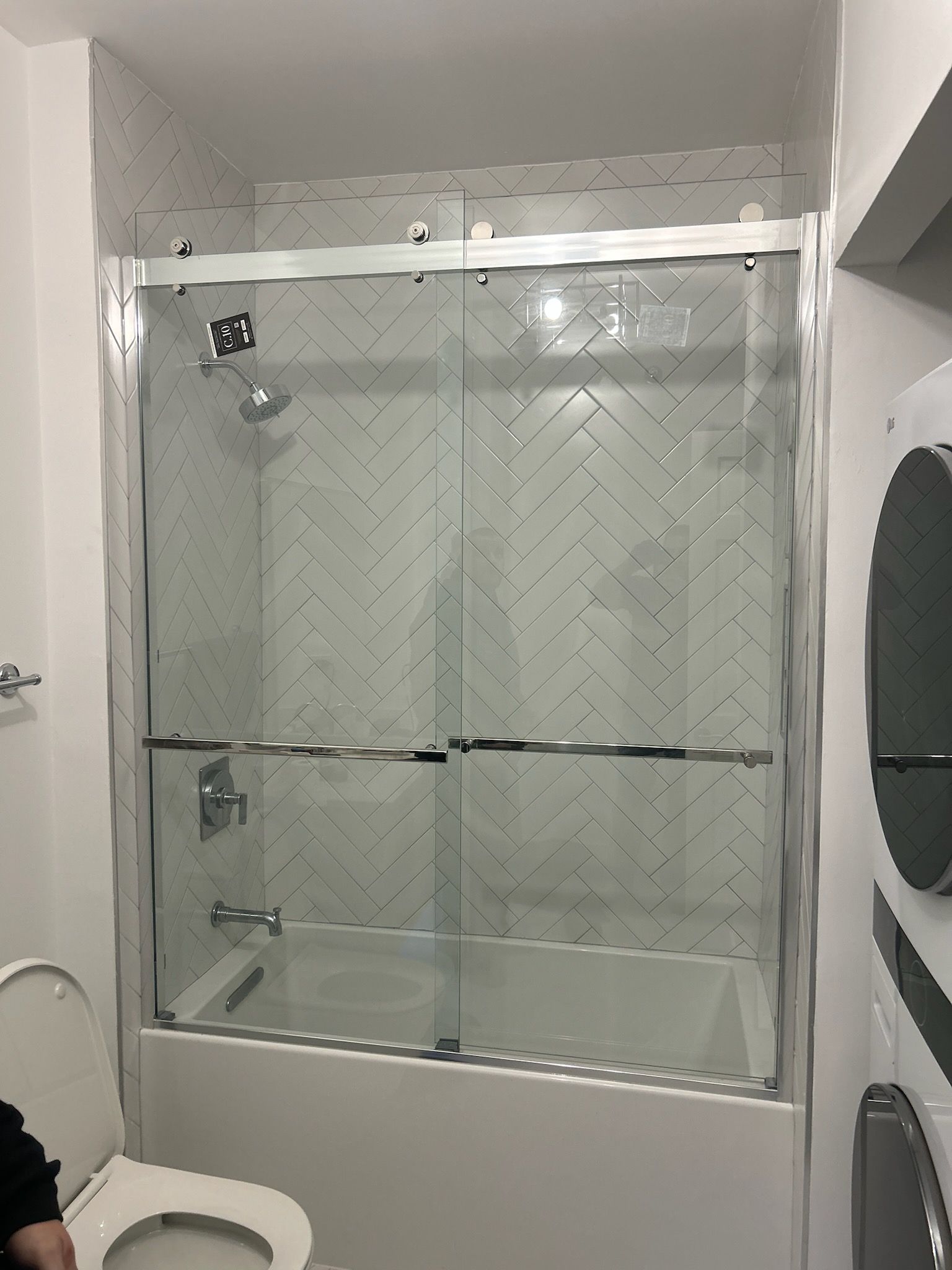 A modern bathroom featuring a white bathtub, chrome sliding glass shower doors, and white herringbone-tiled walls.
