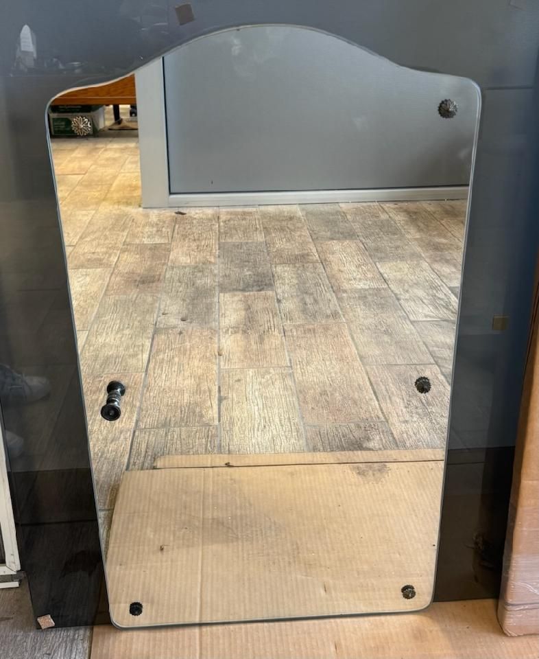 A tall wall mirror with a shaped top, dark reflective border, and visible mounting screws, set against a tiled floor.