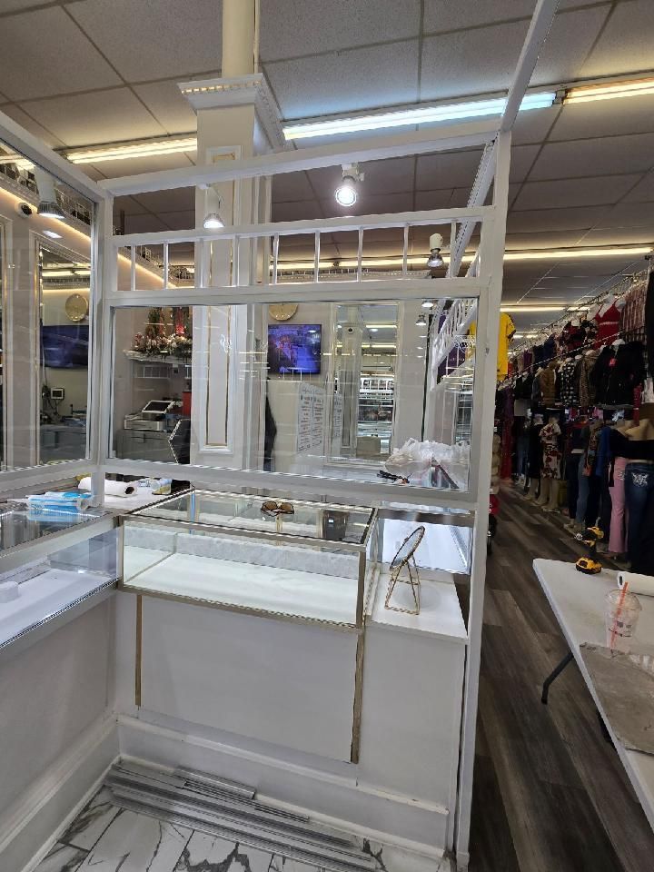 A white display kiosk with a glass showcase inside a retail store, set against a backdrop of hanging clothing racks.