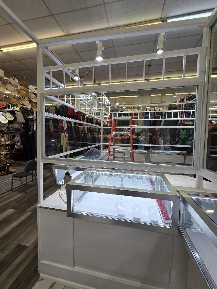 A glass jewelry display case sits in a retail store, with a red stepladder visible through the window structure above.