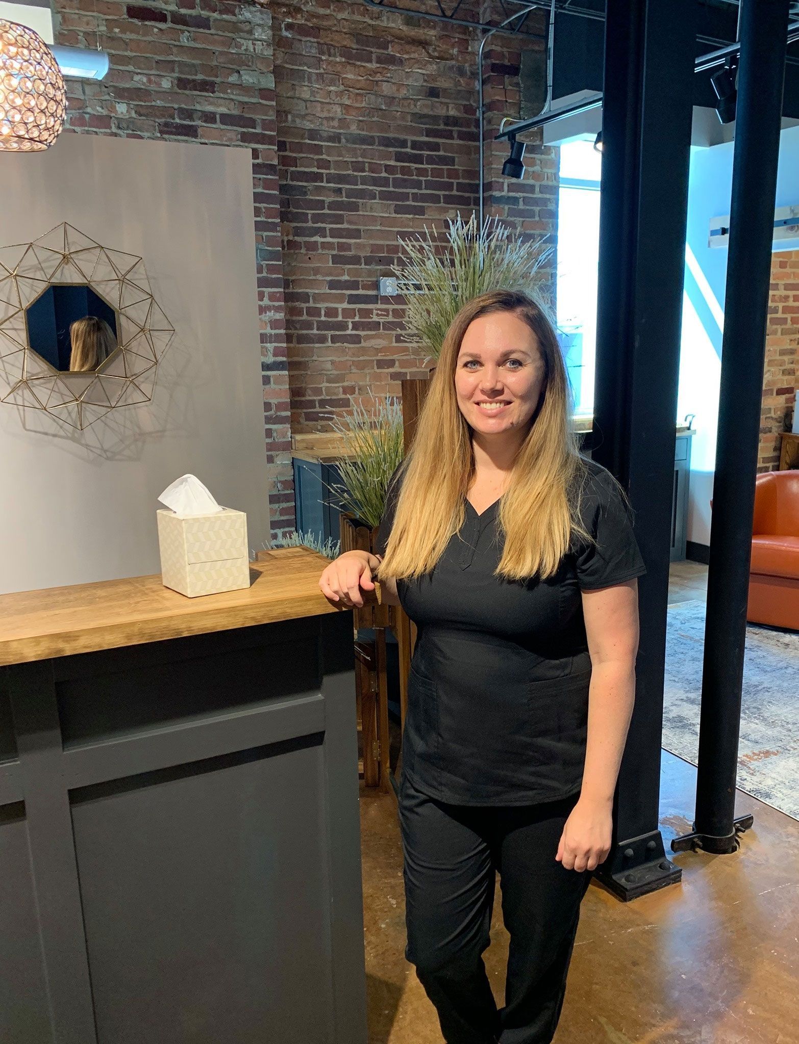 A smiling person with long blonde hair, wearing dark scrubs, stands by a front desk in a brick-walled office space.