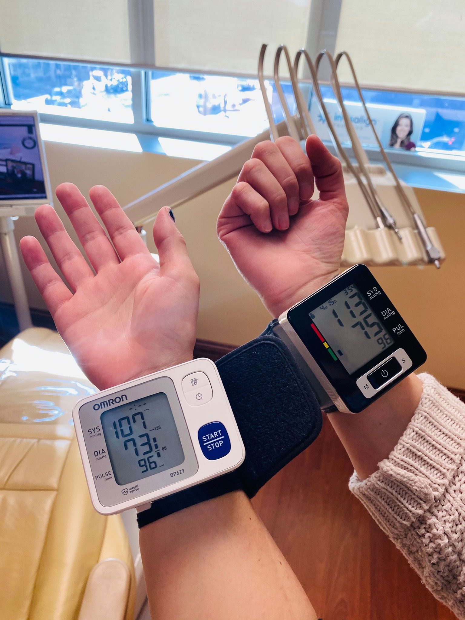 A person's wrists, each wearing a digital blood pressure monitor, in a clinical or dental office setting.