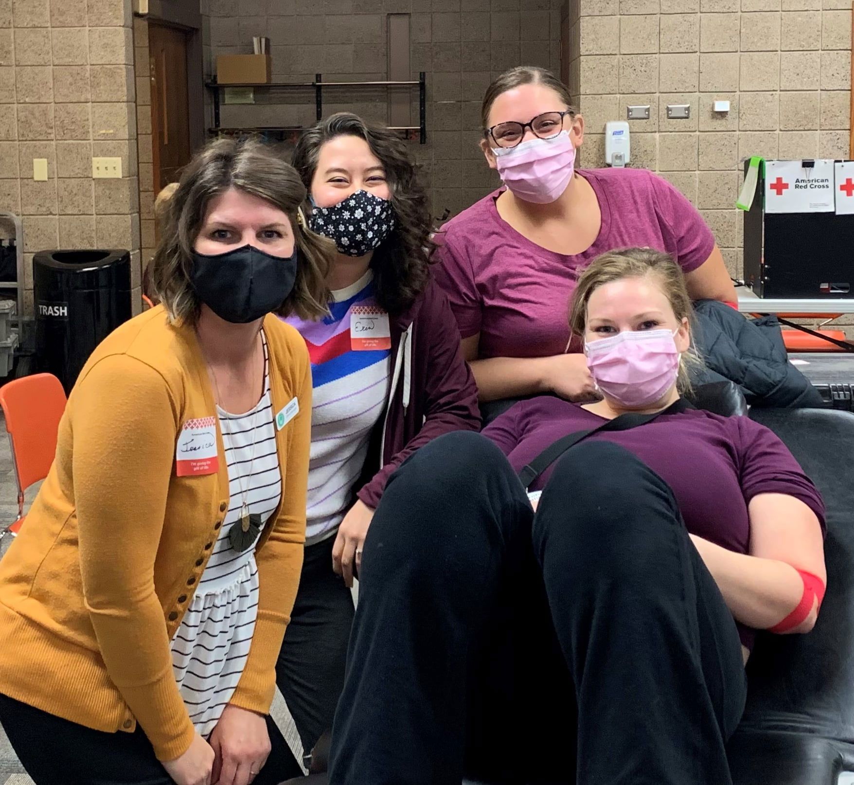Four people wearing face masks pose in a room with a blood drive sign, one sitting in a chair and three standing.