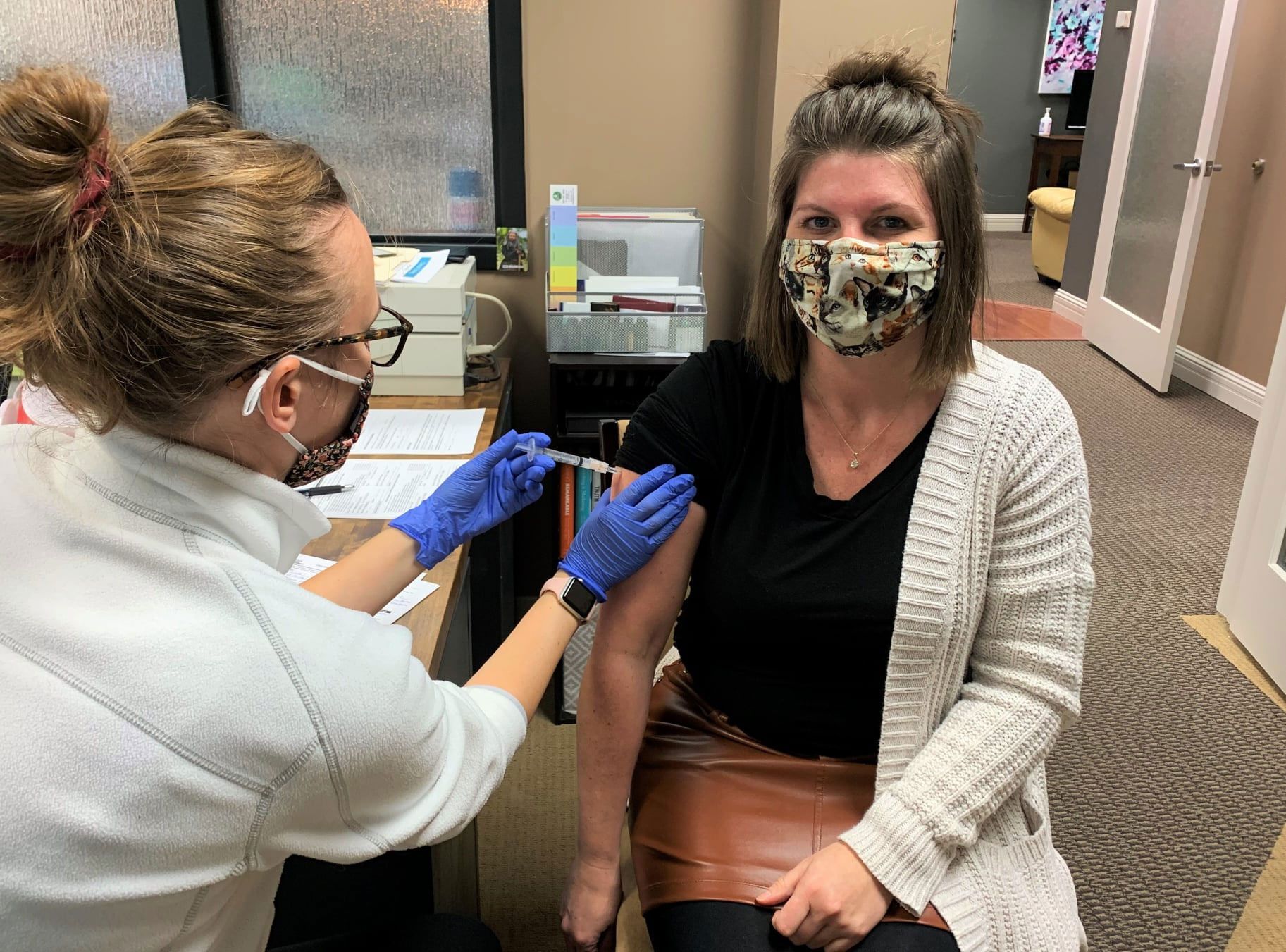A healthcare worker in blue gloves administers a vaccine into the arm of a seated person wearing a patterned face mask.