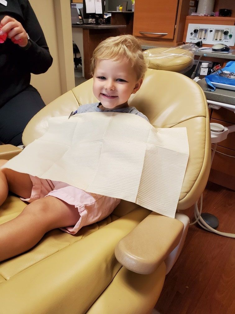 A smiling child sits in a dental chair wearing a protective paper bib.