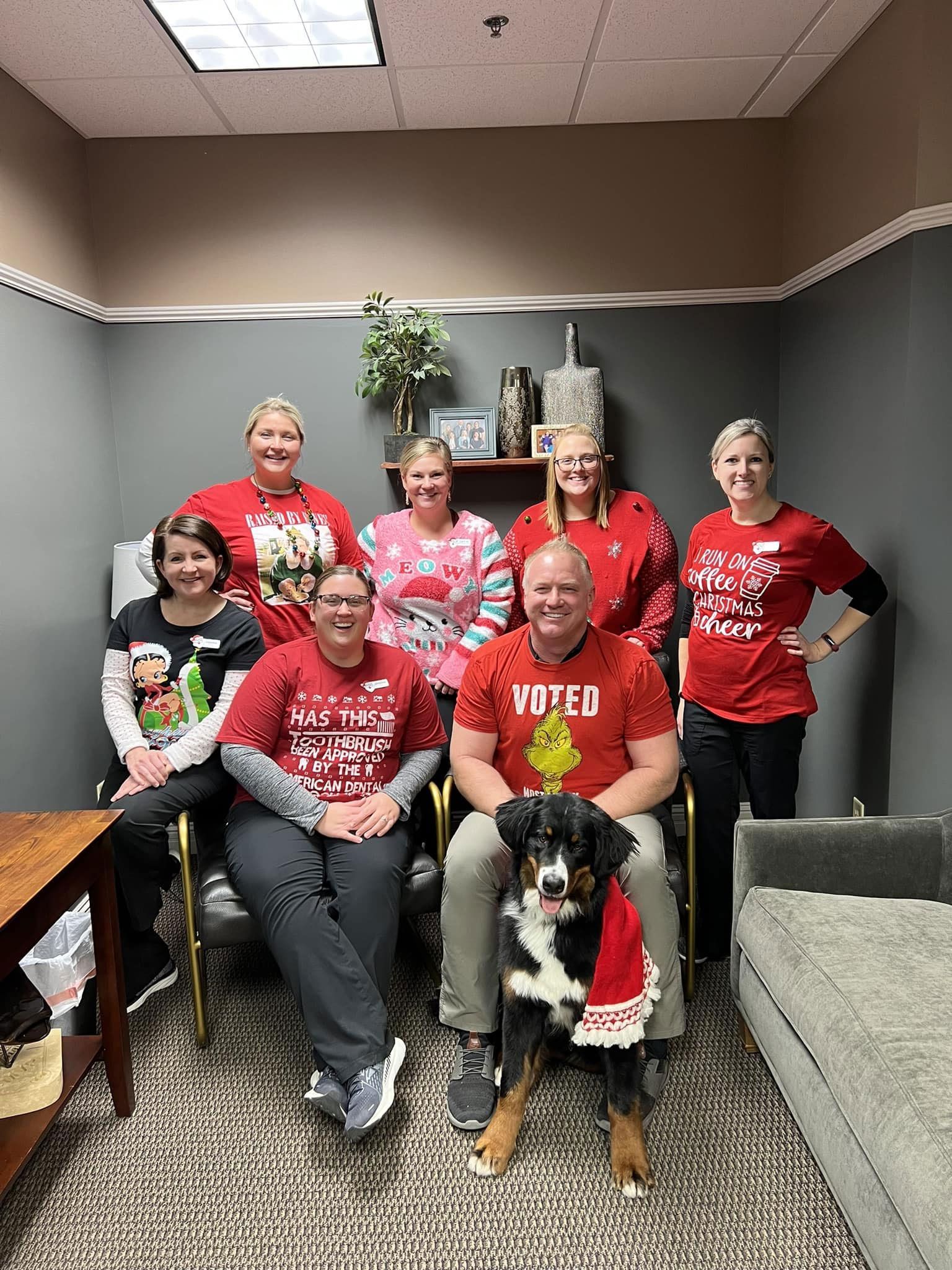 A group of seven people in red holiday-themed clothing poses with a dog wearing a scarf in a decorated office.