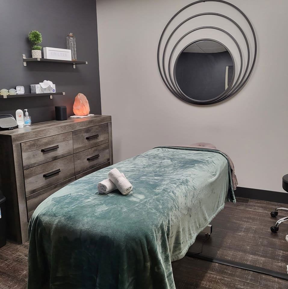 A professional spa treatment room with a massage table, dark wood dresser, wall shelves, and a decorative circular mirror.