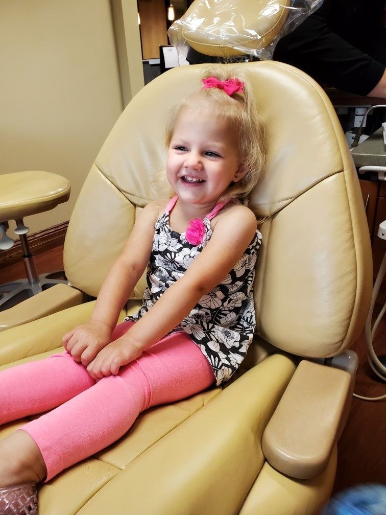 A smiling child sits in a tan dental chair wearing a black and white floral top and pink pants.