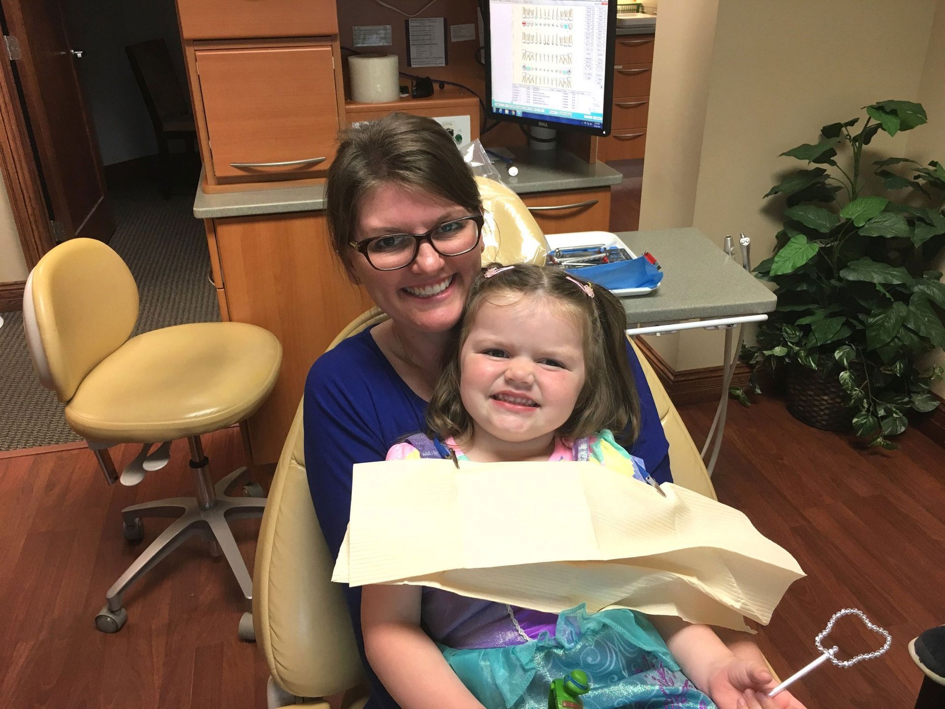 An adult and child sit in a dentist’s chair, both smiling, with a protective bib placed over the child.